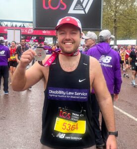 This is a picture of a caucasian man wearing a red and white hat and a black Nike vest top. He is holding his medal out after running the London Marathon.