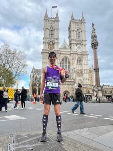 This is an image of a thin man wearing a purple vest top and knee high black socks. He is holding out his medal after running the London marathon. There is a cathedral in the background. 